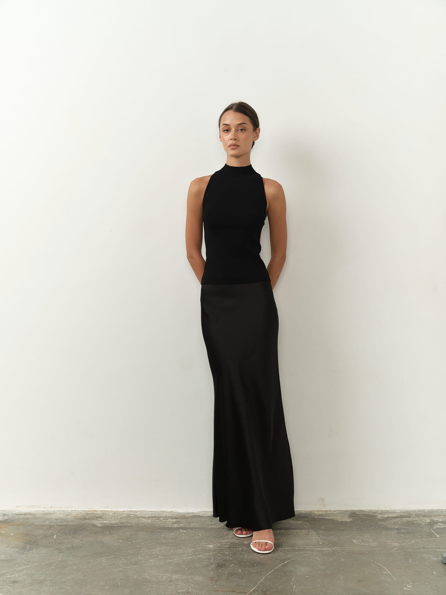 Sleek sleeveless black top featuring a high neck, fitted silhouette, and lightweight fabric, paired with a black satin skirt for a chic and minimalist aesthetic.