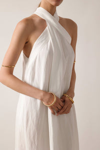  A woman in a white linen maxi Melina dress from NÓRA, complemented by gold bracelets, epitomizing grace and sophistication.