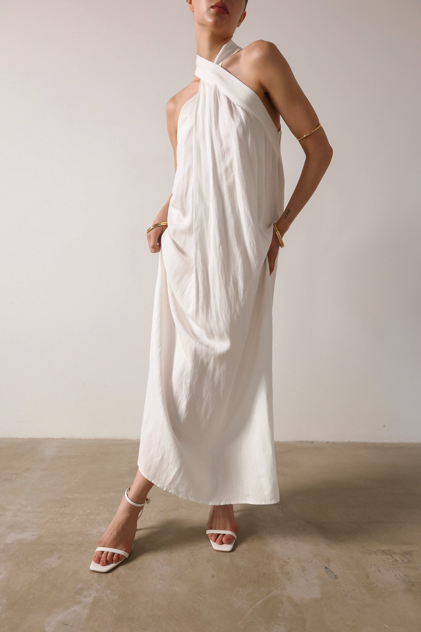  A woman in a white linen maxi Melina dress from NÓRA, complemented by gold bracelets, epitomizing grace and sophistication.