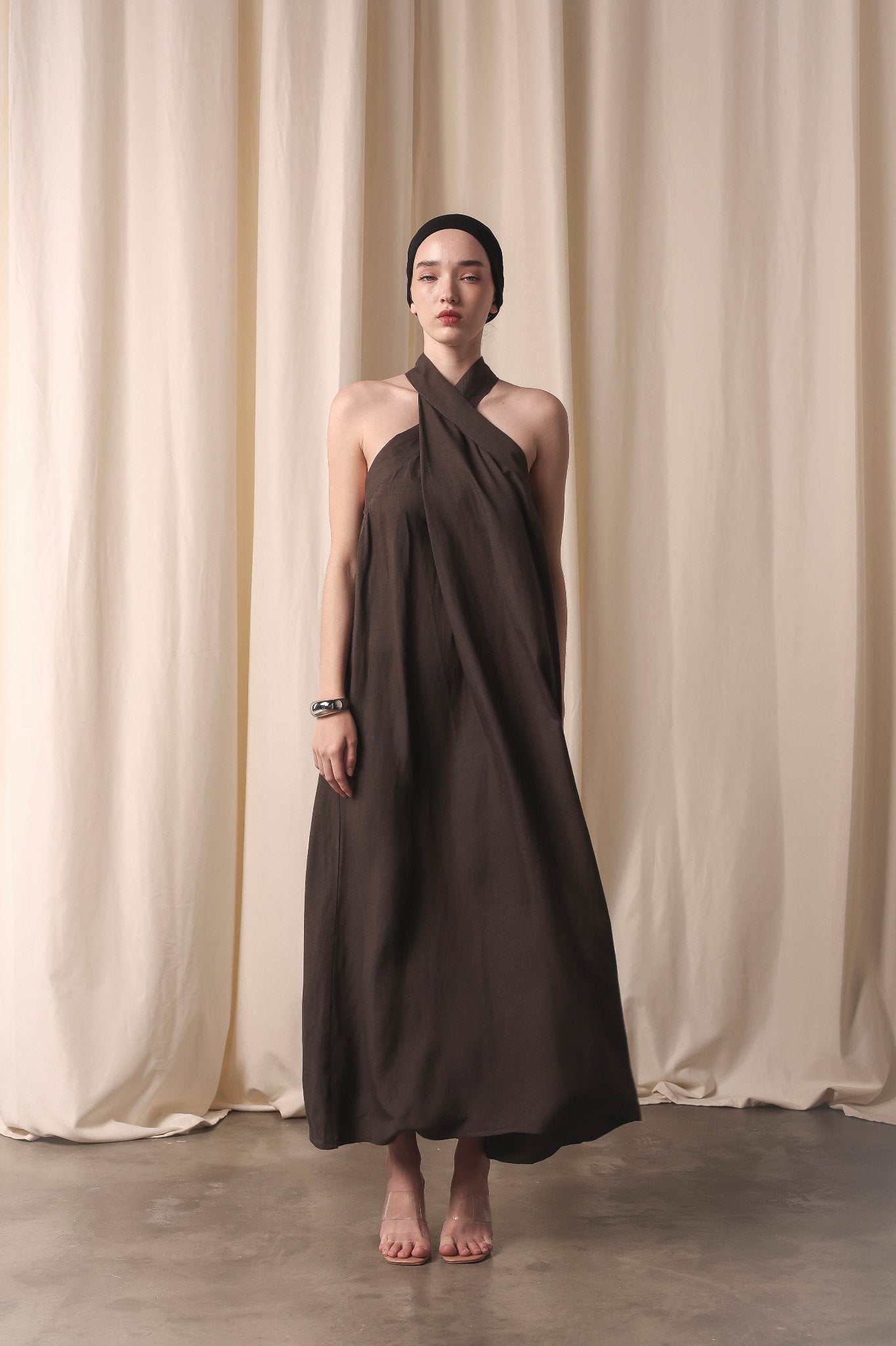 A woman models the Mélina linen maxi dress in brown, standing poised in front of a curtain, exuding sophistication.