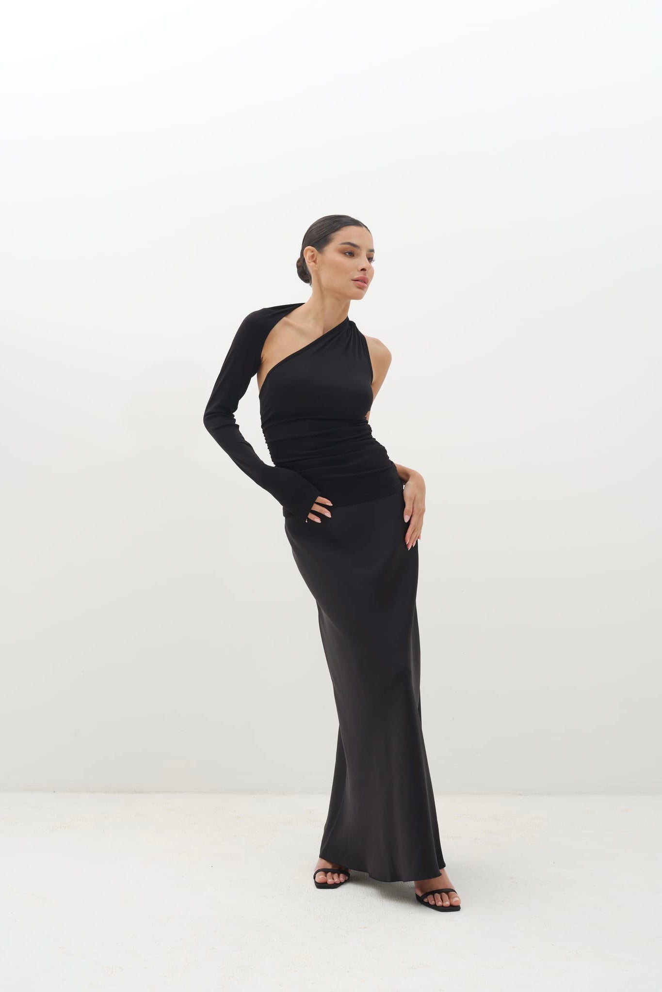 Woman wearing a black one-shoulder top with a long sleeve, paired with a floor-length black skirt.