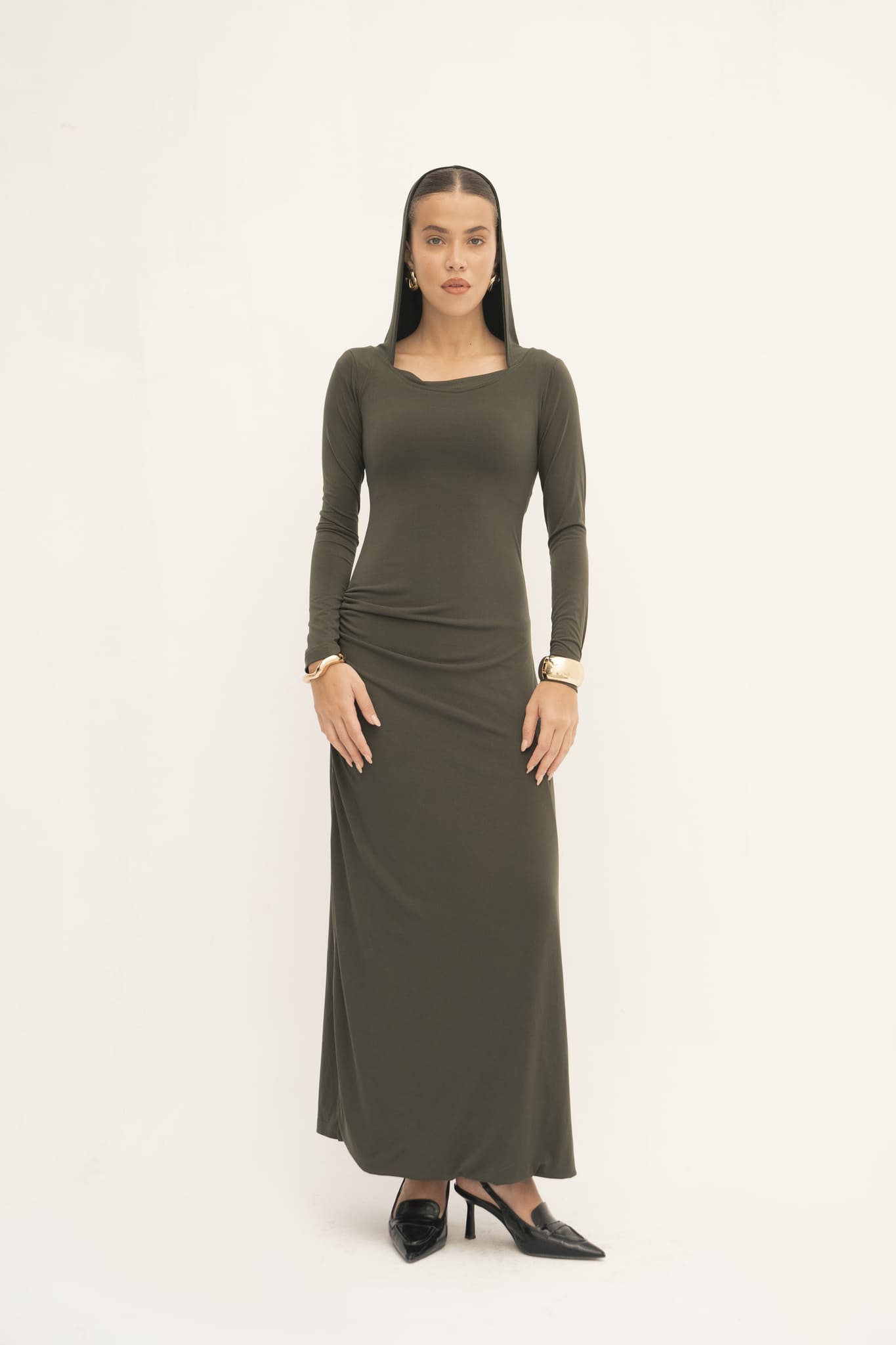 A woman stands against a plain backdrop, showcasing the dark green Olivia Dress with long sleeves. The dress's floor-length design enhances her straight long hair, and she accessorizes elegantly with hoop earrings and bracelets. Her look is finished with black pointed-toe heels.