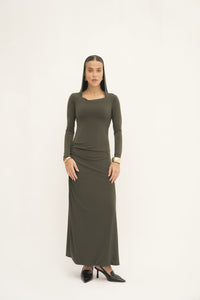 A person poses confidently against a simple backdrop, highlighting the sophisticated floor-length design of the Olivia Dress in dark green. Their black pointed shoes enhance the outfit, and their long straight hair paired with gold hoop earrings contribute to their polished look.