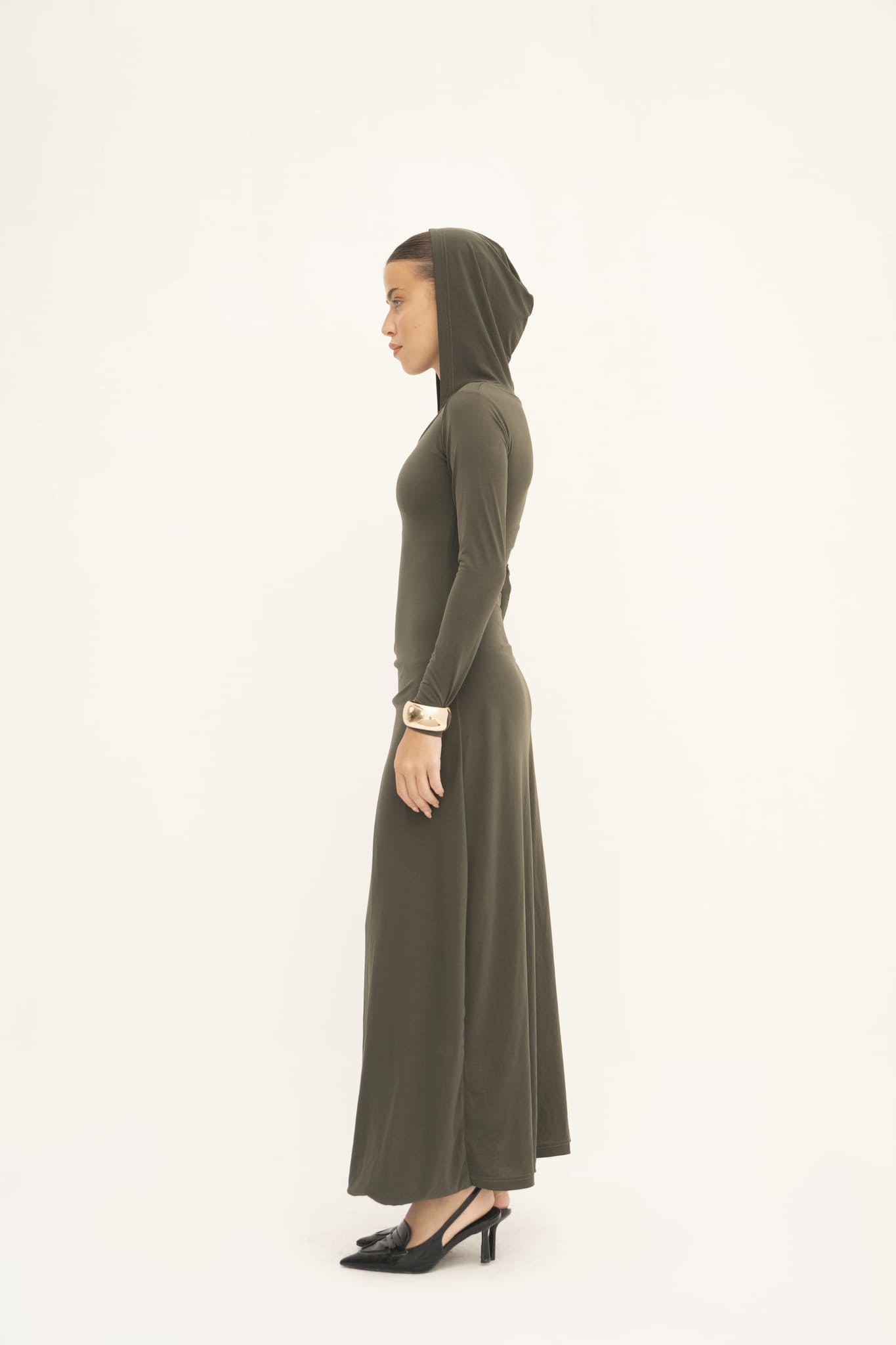 A woman is seen in profile against a white background, showcasing the Olivia Dress—a floor-length design in olive green with a hood. Made from jersey knit fabric, she complements it with black heels and a gold bracelet adorning her wrist.