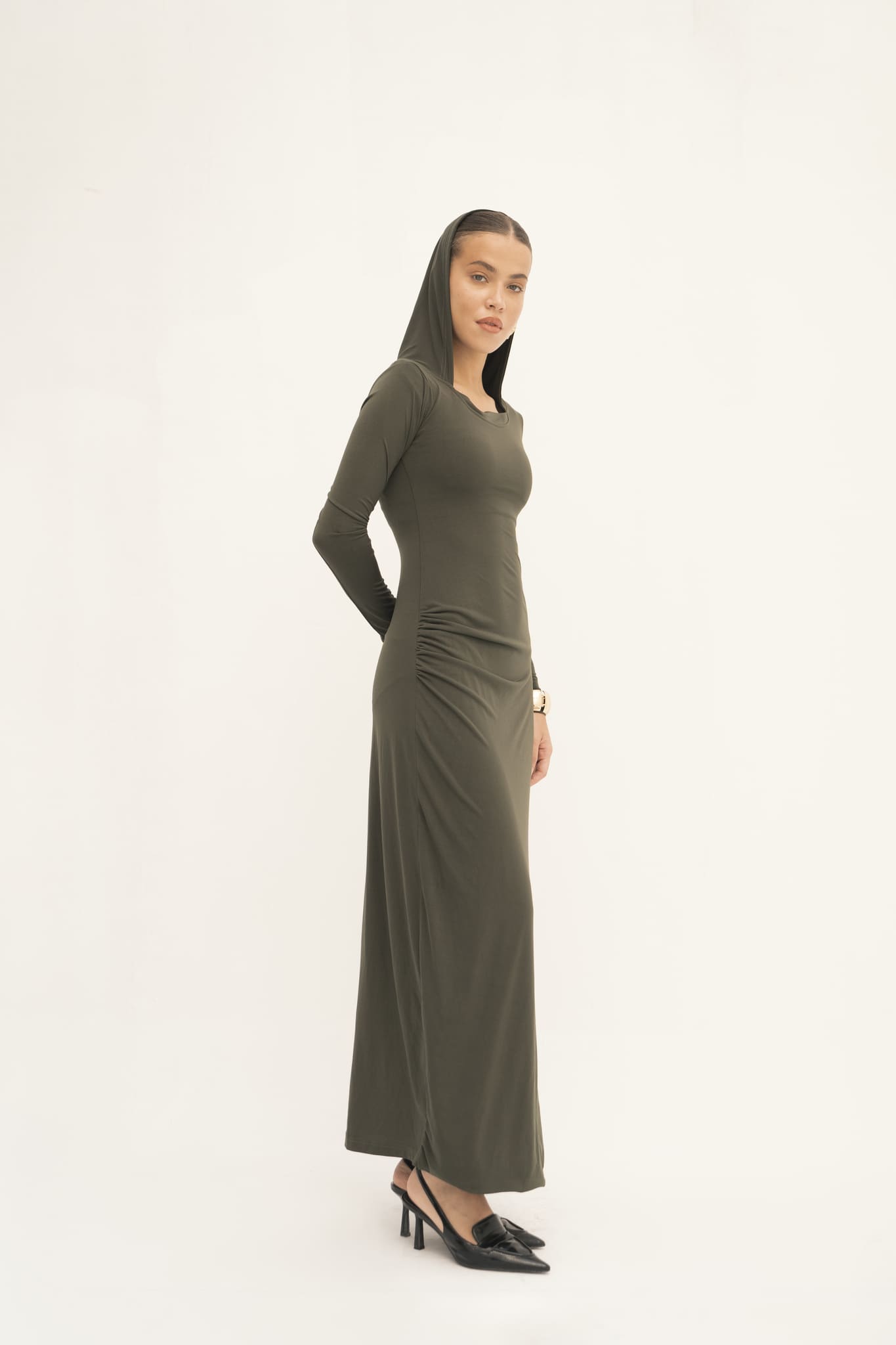 A woman dressed in an olive green Olivia Dress crafted from jersey knit fabric pairs it with black heels as she stands against a light background. Her gaze is forward with a relaxed expression, and her dark hair is styled under a head covering.