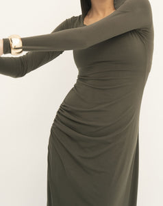 A person wearing the long-sleeved Olivia Dress in olive green, featuring a fitted silhouette and crafted from soft jersey knit fabric with a ruched detail on the side. The image showcases the upper body and intricate dress details, complemented by a gold bracelet set against a plain white background.