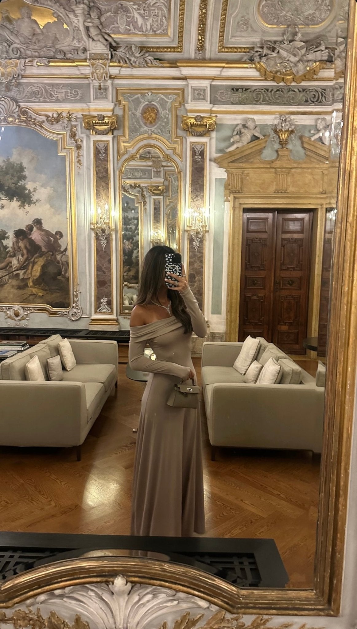 A woman with long, wavy dark hair stands beside a light-colored couch, facing away from the camera. She is wearing an elegant Otis dress made of high-quality spandex fabric. The form-fitting pale grey gown flows effortlessly to the floor.