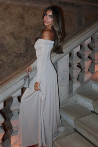 Woman in an elegant off-the-shoulder, long-sleeve beige dress, standing on a staircase. She is holding a small gray handbag and looking over her shoulder with a smile. The setting appears luxurious, with marble textures on the walls and classic stone banisters.