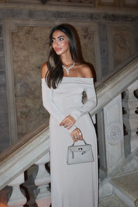 Woman in an elegant off-the-shoulder, long-sleeve beige dress, standing on a staircase. She is holding a small gray handbag and looking over her shoulder with a smile. The setting appears luxurious, with marble textures on the walls and classic stone banisters.