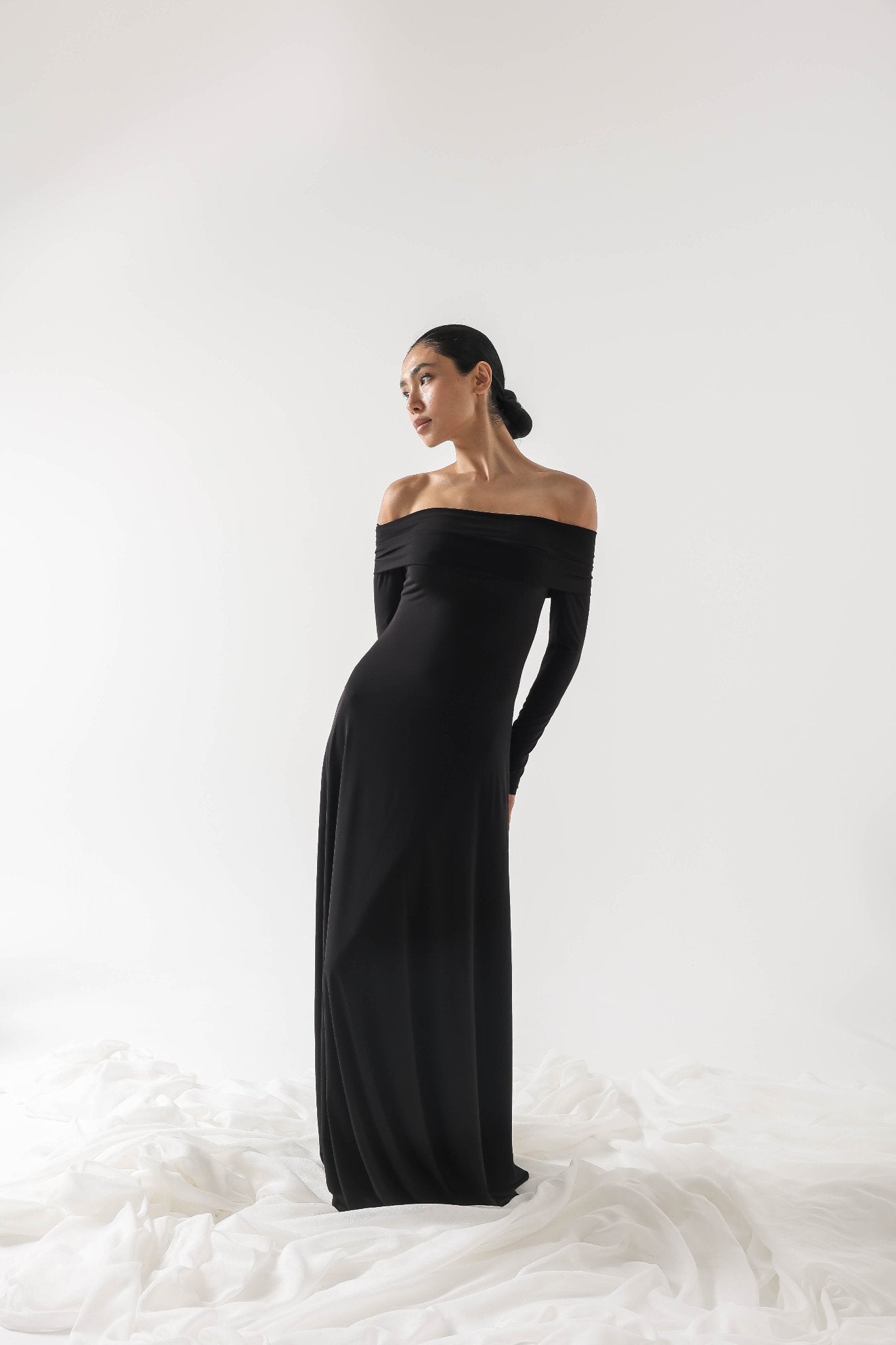 A woman stands gracefully against a white background, wearing an elegant Otis dress made from high-quality spandex fabric. Her dark hair is neatly tied back, and she gazes to her left with a serene expression. The floor is draped with white fabric, contrasting with her dress.