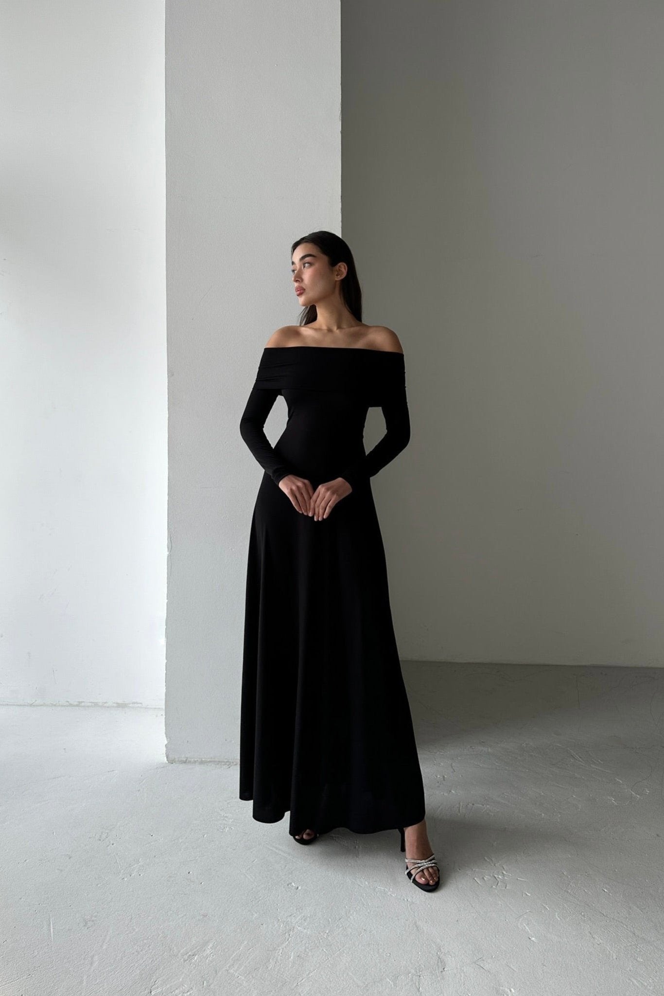 A woman stands in a minimalist, white room, wearing the Otis dress crafted from high-quality spandex fabric. She looks to her left, arms relaxed at her sides, one foot slightly ahead of the other.