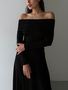 A woman wearing the Otis dress stands with her arms crossed over her body. This elegant maxi dress, crafted from high-quality spandex fabric, features long sleeves and a fitted silhouette. The background is a plain, light-colored wall, and her face is slightly turned to the side.