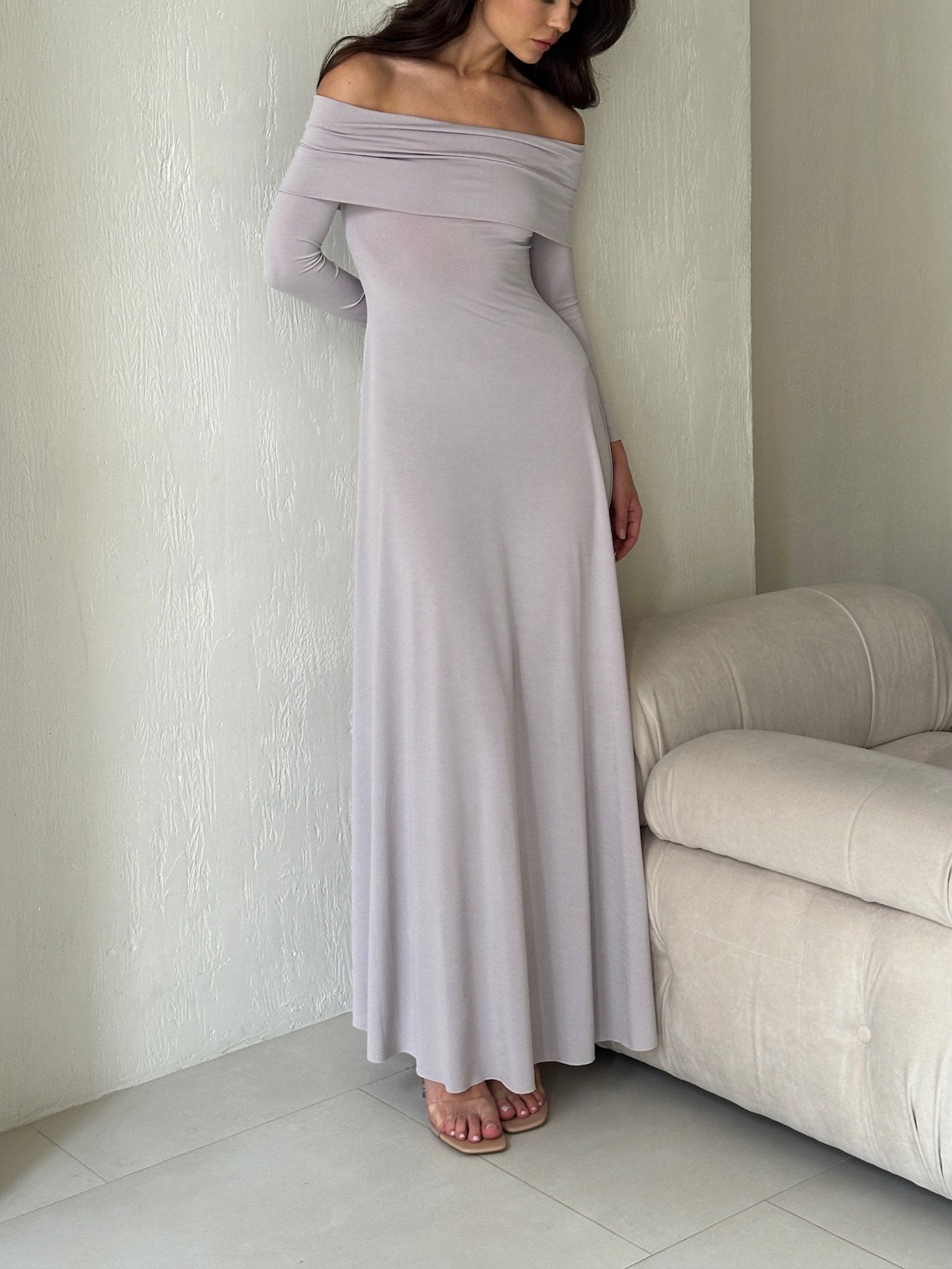 A woman wearing an Otis dress in light grey is standing beside a beige couch. The elegant maxi dress features long sleeves and flows down to her ankles, made from high-quality spandex fabric. She stands against a white textured wall, and the floor is tiled in light grey.