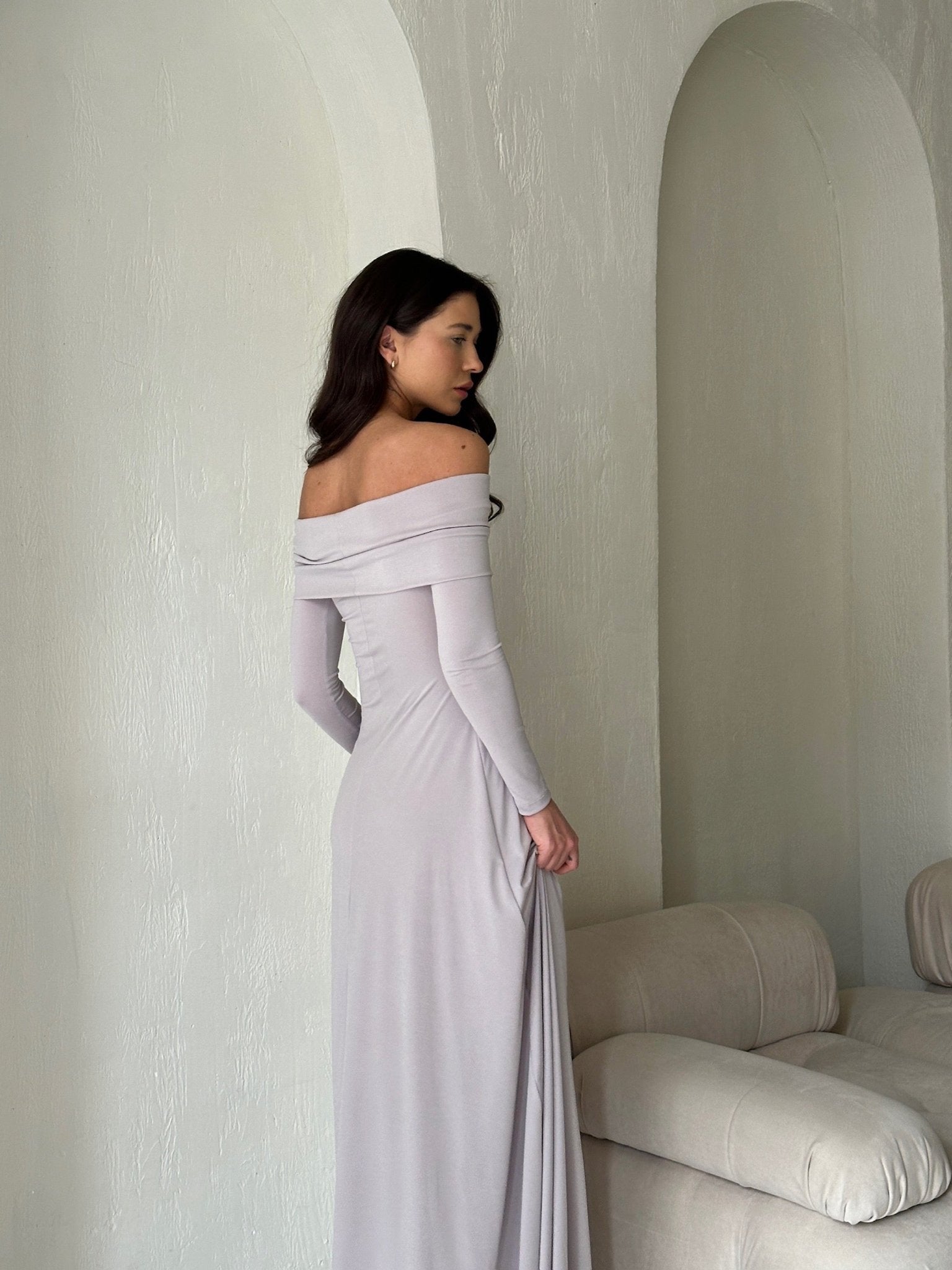 A woman with long, wavy dark hair stands beside a light-colored couch, facing away from the camera. She is wearing an elegant Otis dress made of high-quality spandex fabric. The form-fitting pale grey gown flows effortlessly to the floor against off-white textured walls.