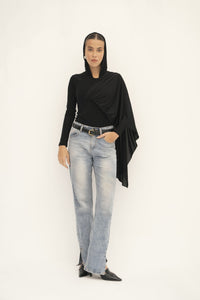 A person dons a black Paulina Top, featuring an asymmetrical drape, paired with light blue jeans. Against a plain backdrop, they strike a pose with one hand in their pocket and wear pointed black shoes, embodying effortless style.