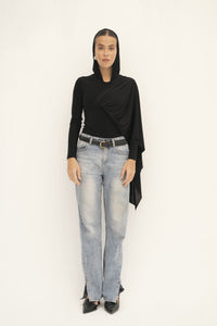 A person poses against a plain background wearing a black headscarf and the Paulina Top, crafted from luxurious fabric with an asymmetrical drape. They pair this look with light blue jeans and black pointed shoes, maintaining a neutral expression.