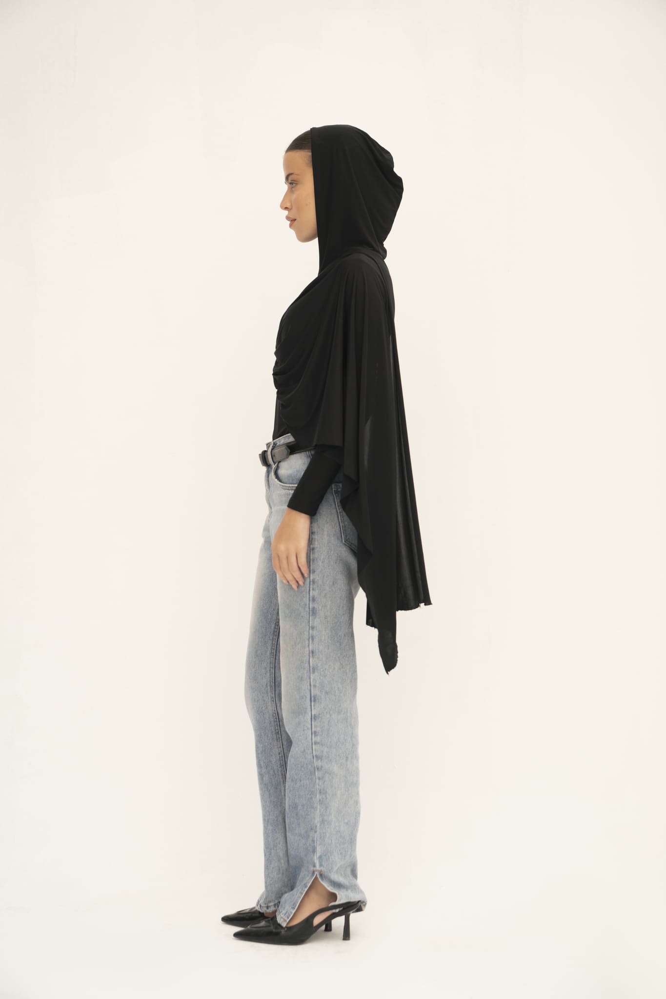 A woman stands in profile against a plain background, showcasing the Paulina Top in black, featuring an asymmetrical drape. She pairs it with light blue jeans and black pointed heels, highlighting the luxurious fabric that enhances the modern and minimalistic aesthetic.