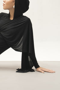 A person wearing the Paulina Top, an elegantly draped black hooded garment, with one arm resting gracefully on the floor. The luxurious fabric's flowing sleeves add elegance to the minimalistic setting, accented by a light-colored backdrop. The face remains partially visible, showcasing sophisticated style.