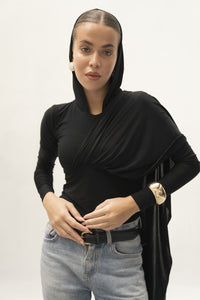 A person with a confident expression wears the Paulina Top in black and light blue jeans, accentuating their look with a large gold bracelet and white earring. The ensemble exudes elegance, enhanced by the luxurious fabric and subtle asymmetrical drape against the plain white background.