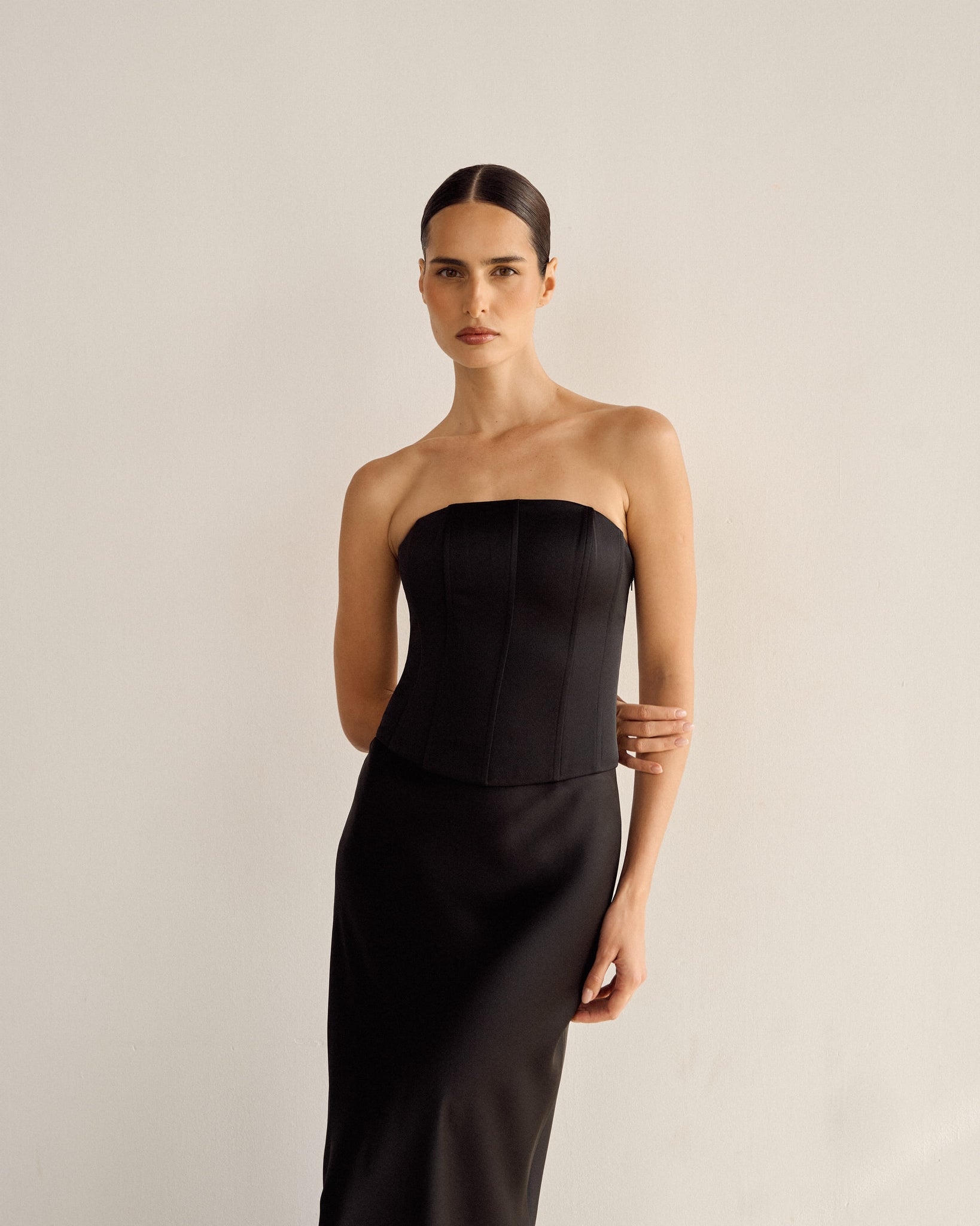 A person with long dark hair is wearing the Regina Corset Set in Black, featuring a strapless satin top and matching maxi skirt, posing against a plain white background. One arm is placed behind their back, echoing the elegance of the ensemble's Bridgerton-esque design, while the other rests on their hip.