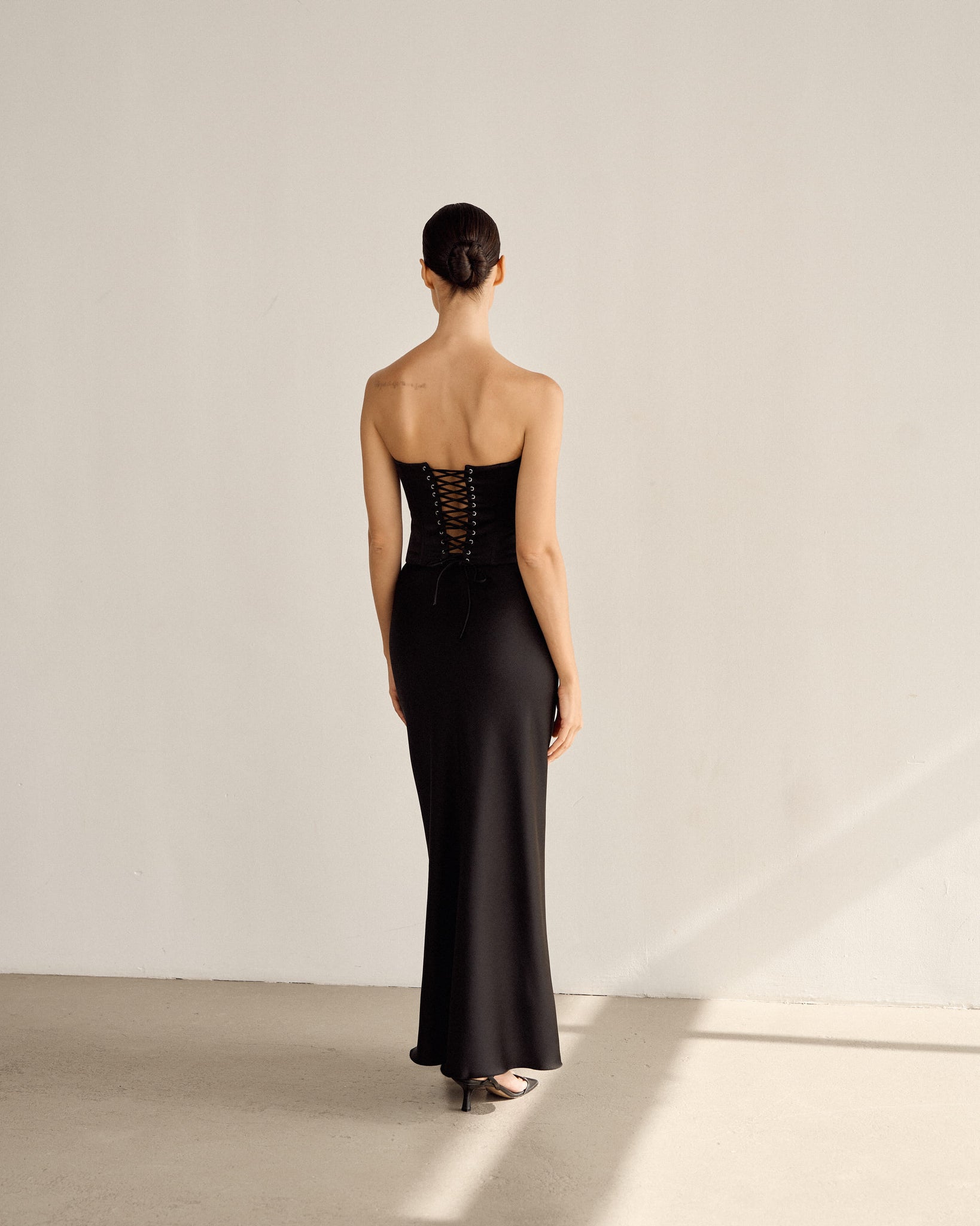 With their back to the camera, someone with short hair wears the Regina Corset Set in black, a long strapless dress characterized by a corset top reminiscent of Bridgerton and intricate lace-up details. The soft lighting in the neutral-colored room casts minimal shadows, accentuating the elegant satin fabric of the ensemble.