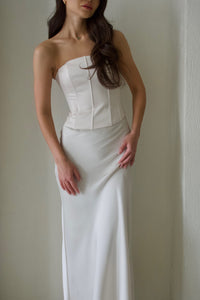 A woman in a strapless Regina corset top, reminiscent of Bridgerton-esque elegance, stands against a plain background. The fitted bodice and sculpting satin fabric enhance her graceful silhouette. Her long, wavy hair cascades over her shoulders as she gently holds the flowing skirt with one hand.