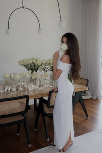 A woman with a feminine silhouette and long brown hair stands in an elegant dining room, wearing the Scarlett Dress—a stunning off-the-shoulder gown with an exquisite ruching effect. She is partially turned towards the camera, holding a white rose to her face. The table behind her showcases clusters of white roses in vases.