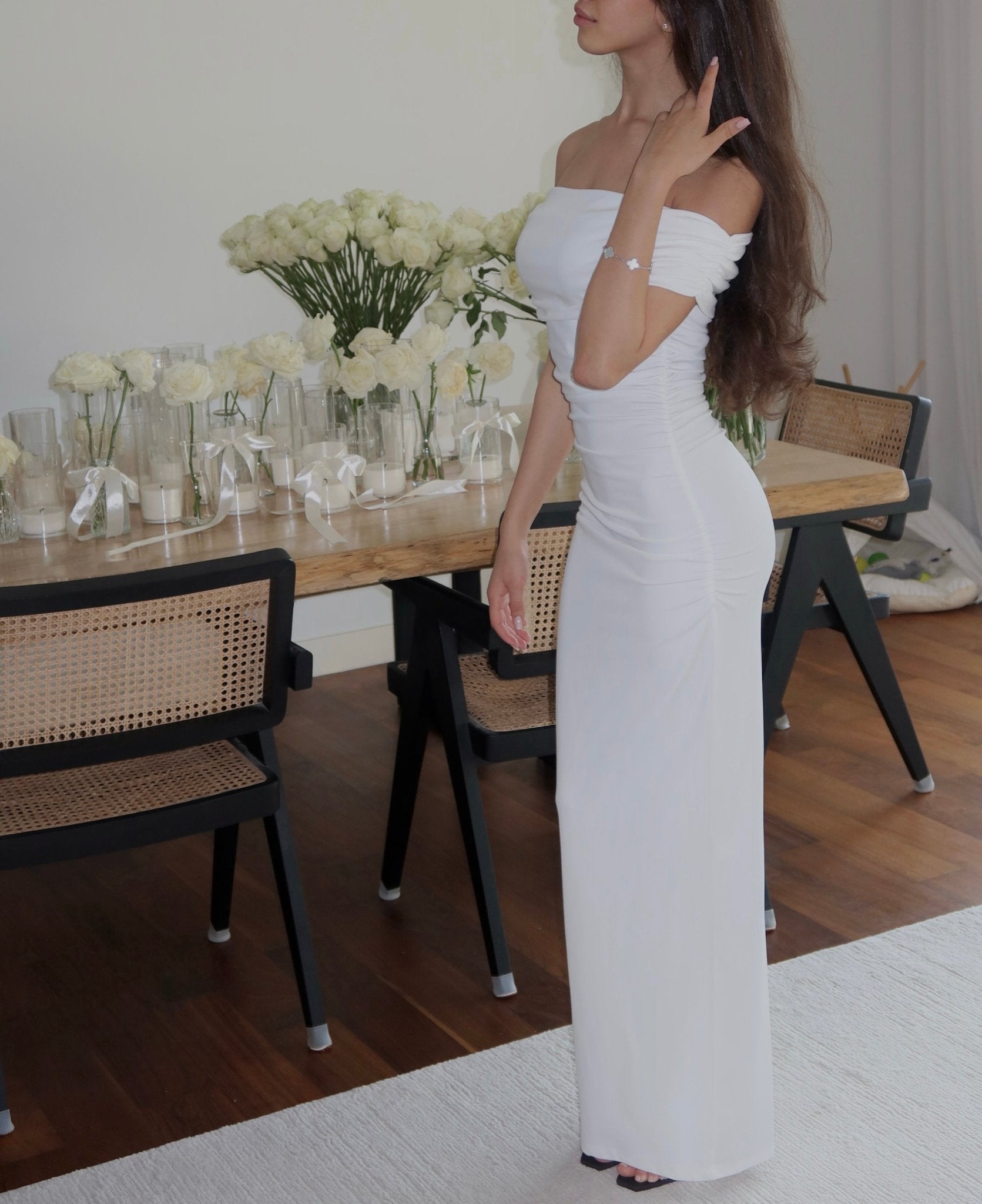 A woman with long brown hair wearing the Scarlett Dress, an off-the-shoulder, floor-length white gown that highlights a feminine silhouette, stands indoors. Behind her is a wooden dining table adorned with vases of white flowers and surrounded by black chairs featuring rattan backs. The wooden floor showcases a partially visible white rug.