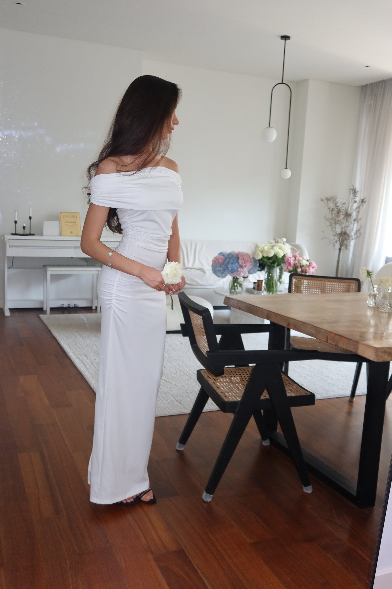 A woman in the Scarlett Dress, featuring a long, off-the-shoulder style with subtle ruching, holds a small bouquet of flowers while standing near a dining table. The room has wooden floors, white walls, and modern furniture, including black chairs around the dining table, and is decorated with vases of flowers.