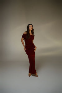 A woman stands confidently, posing in the Scarlett Dress—a maroon off-the-shoulder long gown featuring a stunning ruching effect. She pairs it with shiny heels and her long hair is styled in loose waves, set against a plain, softly lit background.