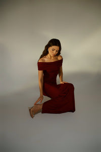 A woman with long dark hair sits on one knee on a light-colored floor. She is wearing the off-the-shoulder, long burgundy Scarlett Dress featuring a ruching effect, paired with clear high-heeled shoes. The background is simple and uncluttered, emphasizing the elegance of her pose and feminine silhouette.