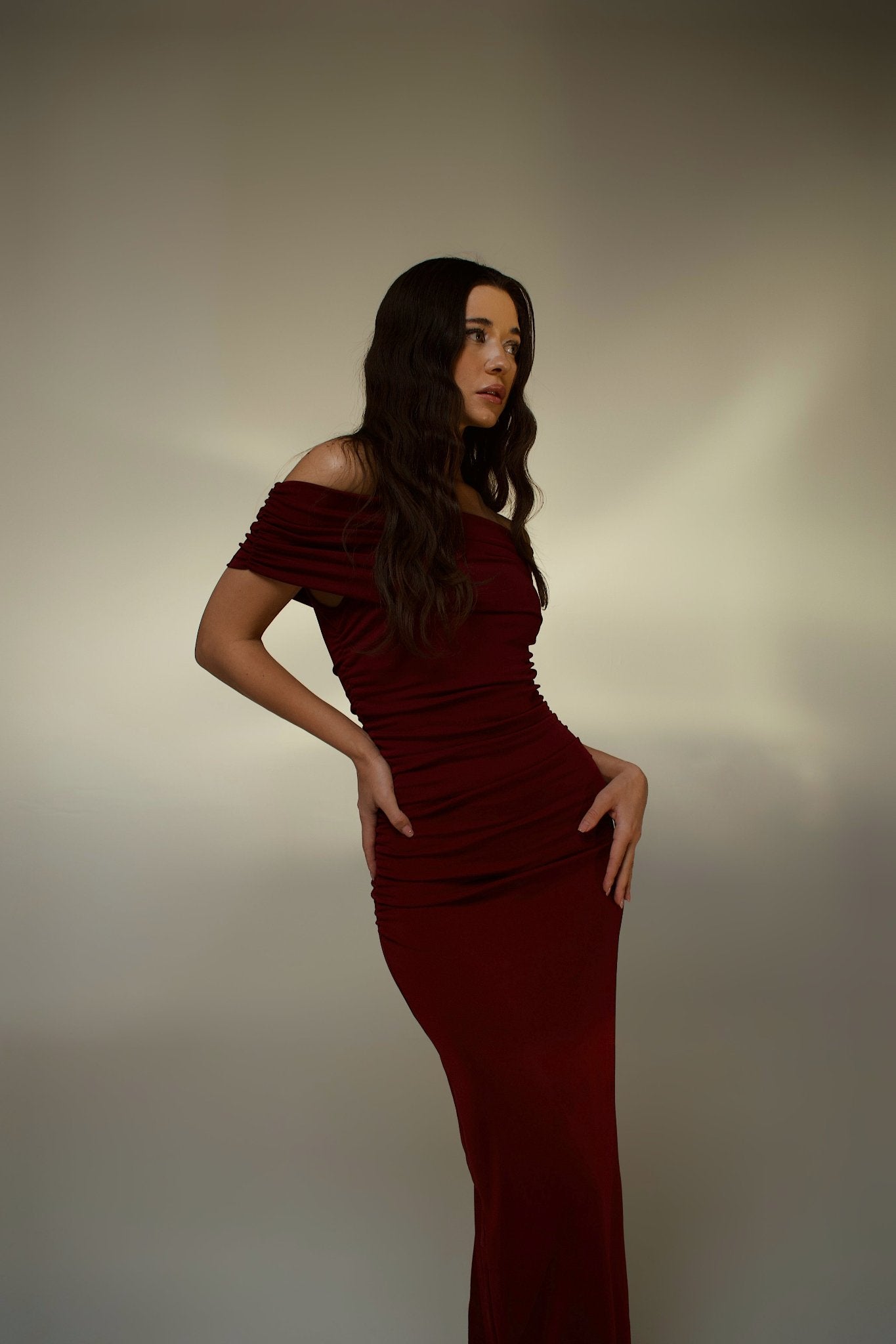 A woman stands against a soft gradient background, wearing the sleek, off-the-shoulder Scarlett Dress in burgundy. Her long dark hair cascades in loose waves, and she gazes to the side with a serene expression, hands resting gracefully on her hips, accentuating her feminine silhouette.