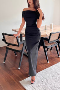 A woman stands in a modern dining room wearing the Scarlett Dress, an off-the-shoulder long gown that highlights her feminine silhouette. She rests one hand on a wooden dining table with black and woven chairs, while her other hand touches her hair. She wears black high-heeled sandals.