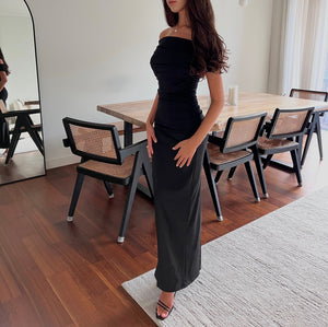 A woman stands in a stylish, modern dining area wearing the elegant Scarlett Dress with its off-the-shoulder design and feminine silhouette, paired effortlessly with high heels. Her long hair cascades freely as she gently rests her hands on her hips. In the background, a wooden dining table accompanied by four chairs and a large mirror completes the scene.