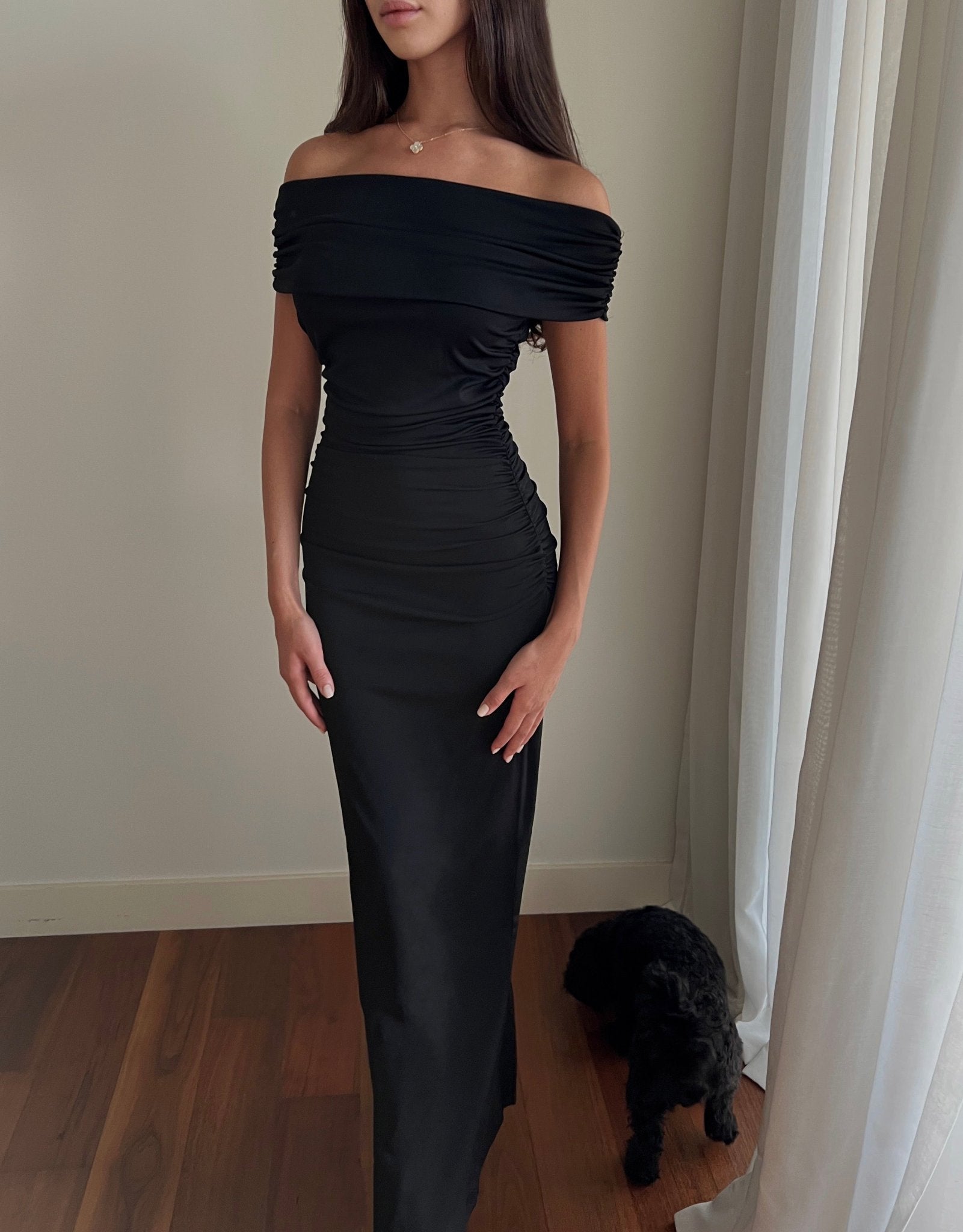 A woman stands in a room wearing the Scarlett Dress, the off-the-shoulder design and ruching effect accentuating her feminine silhouette. She has long, dark hair and is facing slightly to the side. A small black dog sits on the wooden floor near her feet and a white curtain.