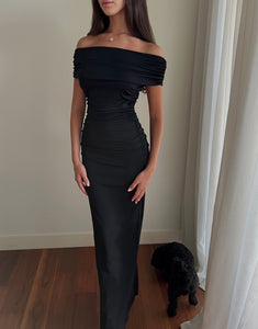 A woman stands in a room wearing the Scarlett Dress, the off-the-shoulder design and ruching effect accentuating her feminine silhouette. She has long, dark hair and is facing slightly to the side. A small black dog sits on the wooden floor near her feet and a white curtain.