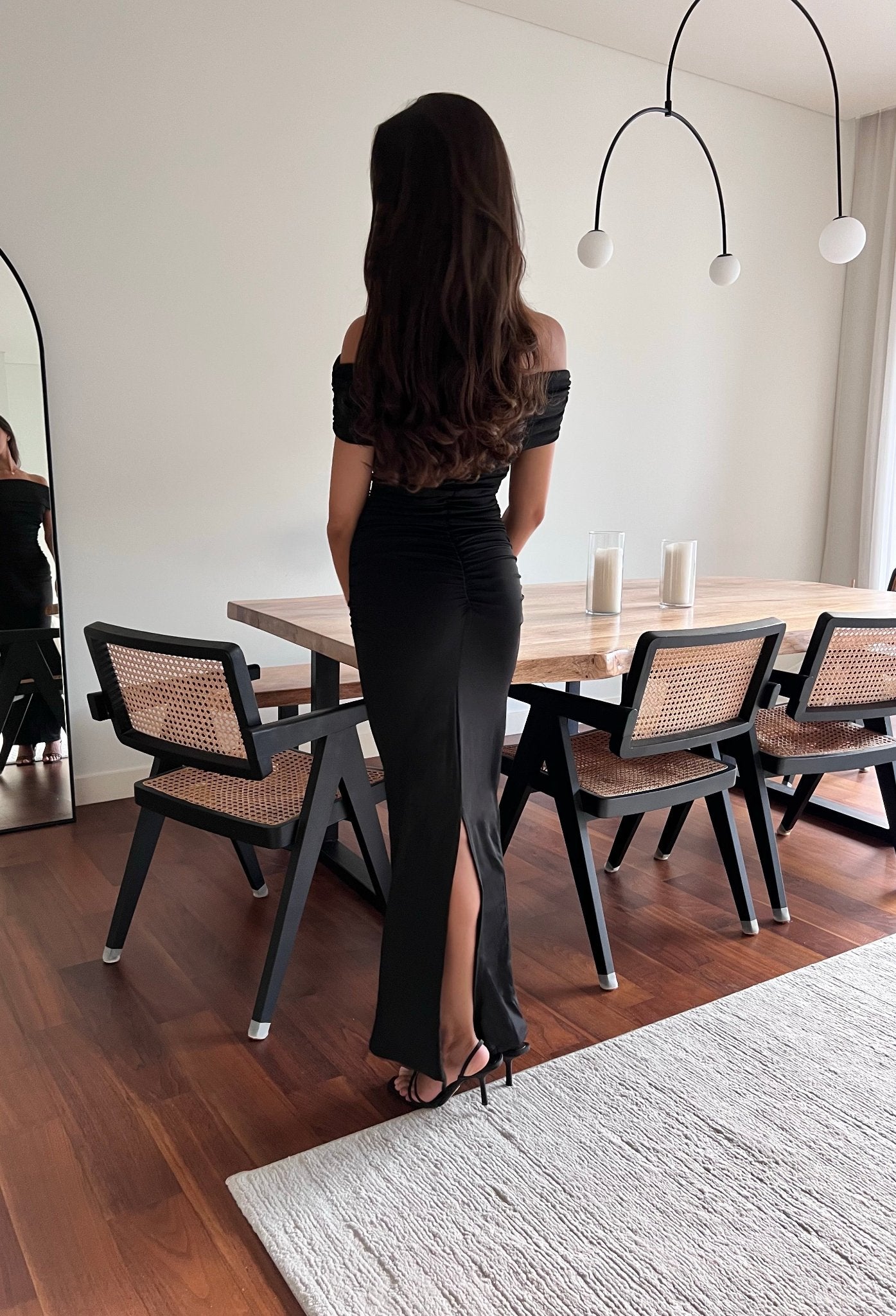 A woman with long, wavy hair stands facing away from the camera, wearing the Scarlett Dress, an off-the-shoulder long gown that accentuates her feminine silhouette. The black floor-length Scarlett Dress features a slit at the back. She stands beside a wooden dining table with cushioned chairs, and a modern light fixture hangs above it.