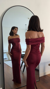 A woman wearing the elegant Scarlett Dress, an off-the-shoulder long gown in maroon, stands in front of a large mirror. With dark shoulder-length hair, she admires her reflection that accentuates her feminine silhouette. The room is adorned with minimalistic décor, featuring white walls and a wooden floor.