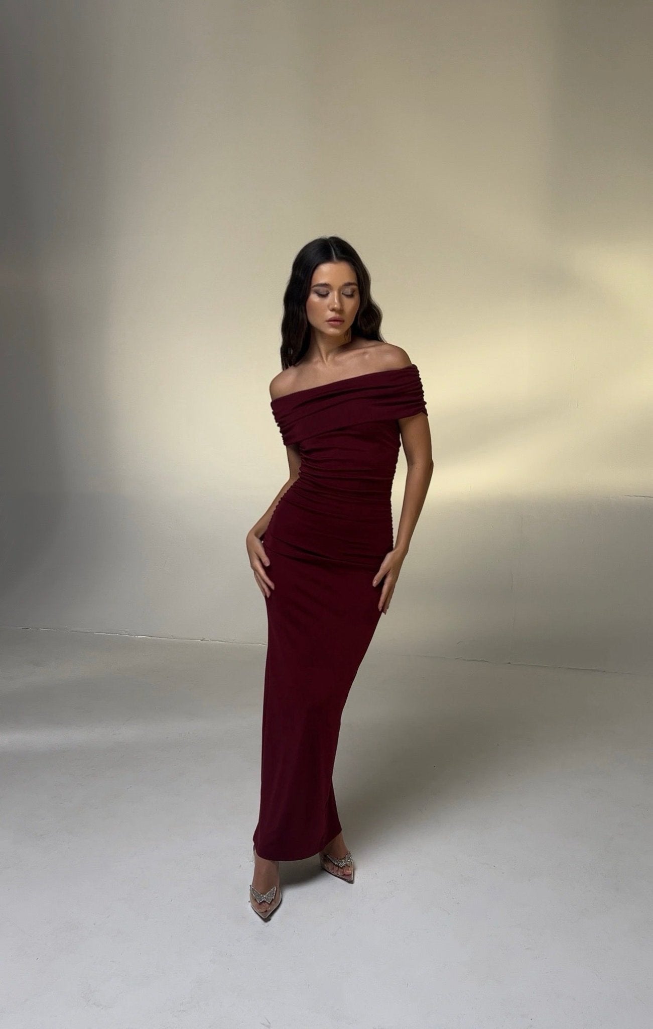 A woman with long dark hair stands in a studio wearing the form-fitting Scarlett Dress, an off-the-shoulder burgundy piece that accentuates her feminine silhouette. The clear high-heeled shoes add elegance to her look, while the softly lit beige-toned background highlights the ruching effect on her dress.