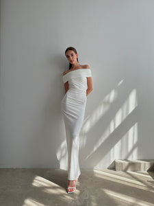 A person stands against a white wall in a well-lit room, wearing the elegant Scarlett Dress. This off-the-shoulder, floor-length gown showcases a fitted feminine silhouette and features a subtle ruching effect. The dress is paired with white open-toe heels while natural light casts shadows on the wall and floor.