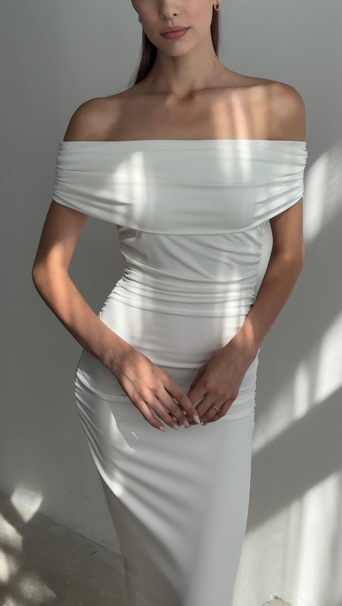 A woman is wearing the Scarlett Dress, an off-the-shoulder long dress that is form-fitting and accentuates her feminine silhouette. She stands with her hands gently clasped in front of her, with light casting soft shadows on both her and the white wall in the background.