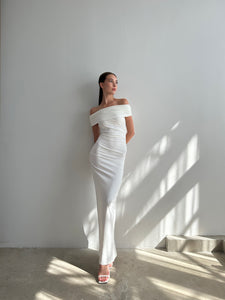 A woman stands against a plain wall with light and shadow patterns, wearing the Scarlett Dress that accentuates her feminine silhouette. Pink high heels add a touch of elegance as she poses with her hands behind her back, gazing to the side. The floor has a polished, concrete finish.