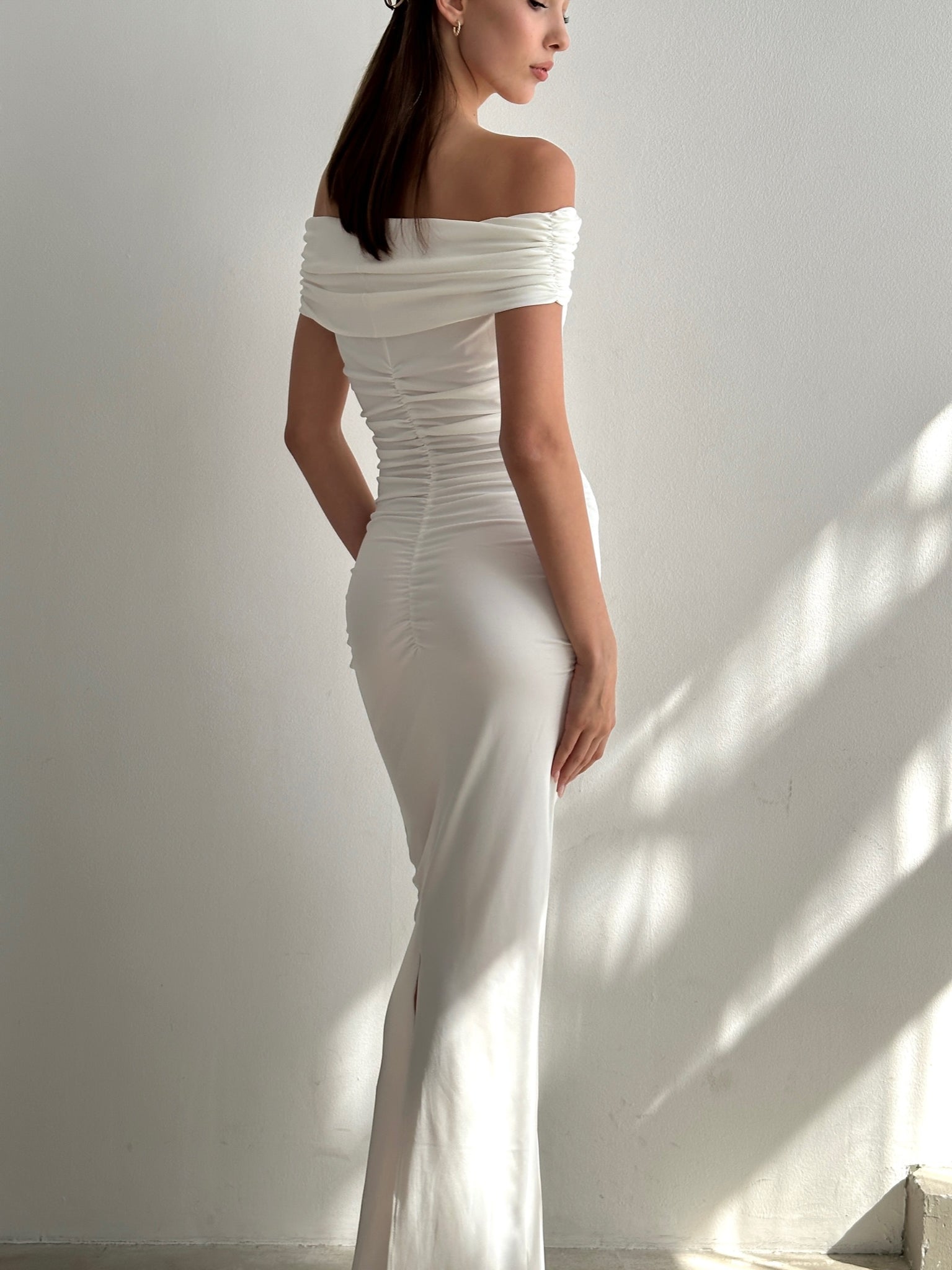 A woman stands against a light-colored wall, wearing the elegant Scarlett Dress, an off-the-shoulder long gown with ruching along the back. Her hair is styled straight and she faces slightly away from the camera, showcasing the feminine silhouette of the dress.