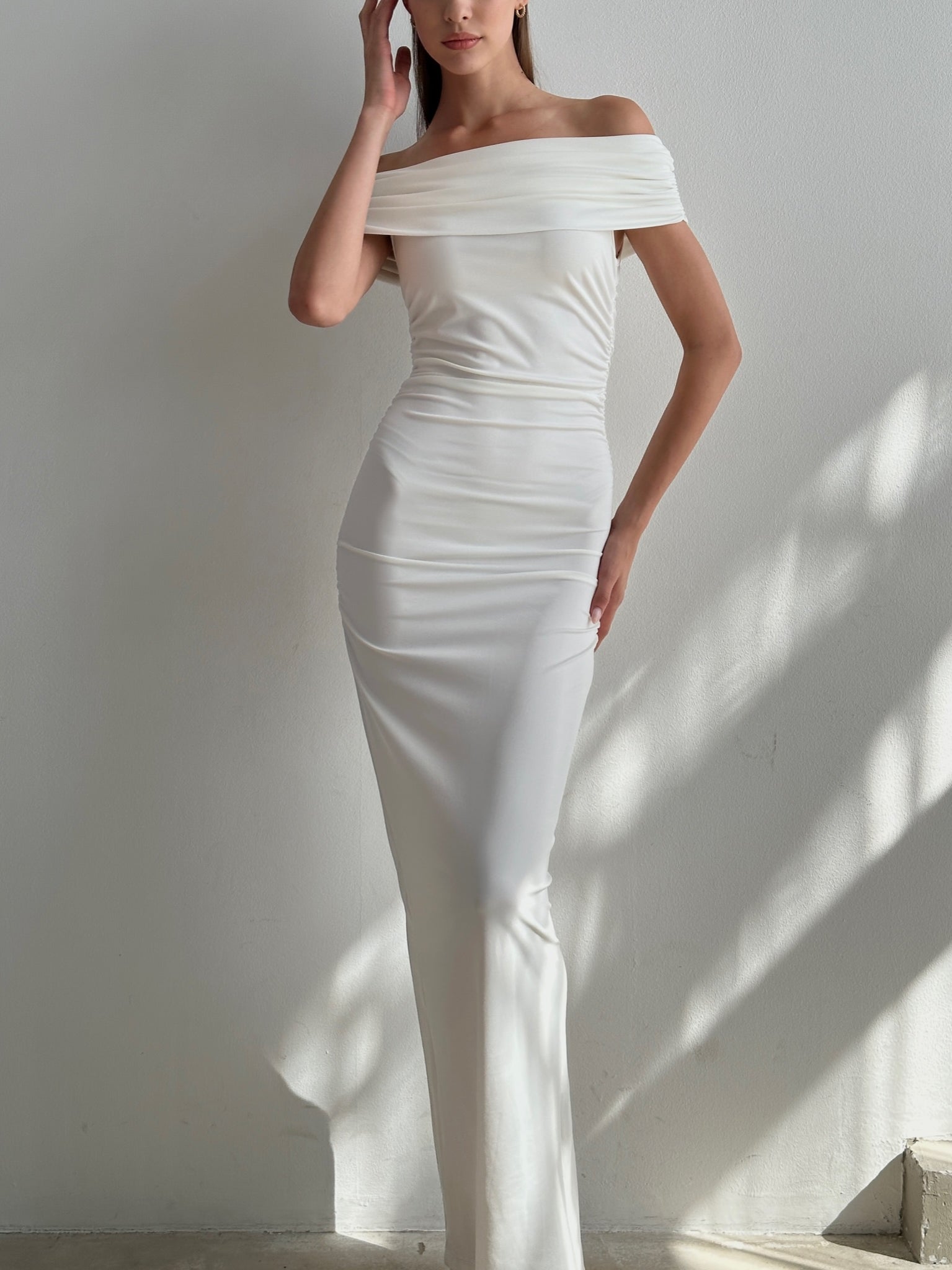 A person poses against a light-colored wall, wearing the elegant Scarlett Dress. The off-the-shoulder long gown features a feminine silhouette and is form-fitting, accentuating the figure with a subtle ruching effect. Soft shadows are cast around them by the lighting as one hand lightly touches their head and the other rests on their hip.