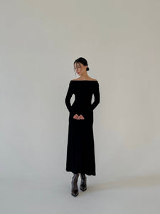 A woman stands against a simple light backdrop, wearing the luxurious fabric of the Sienna maxi dress. The long black dress features off-shoulder and long sleeves, perfectly complementing her shiny metallic ankle boots as she clasps her hands in front and gazes slightly to the side.