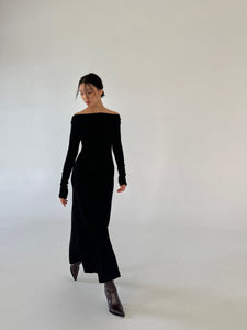 A person with dark hair, wearing a Sienna maxi dress and dark boots, is walking against a plain white background. The versatile dress's luxurious fabric flows gracefully as the person looks down and to the side, with one arm slightly extended.