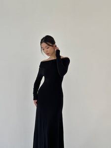 A woman with dark hair styled in a low updo wears the Sienna Maxi Dress, a long-sleeve black gown made of luxurious fabric. She stands against a plain, light-colored background with her face turned slightly downward and one hand lightly touching her hair.