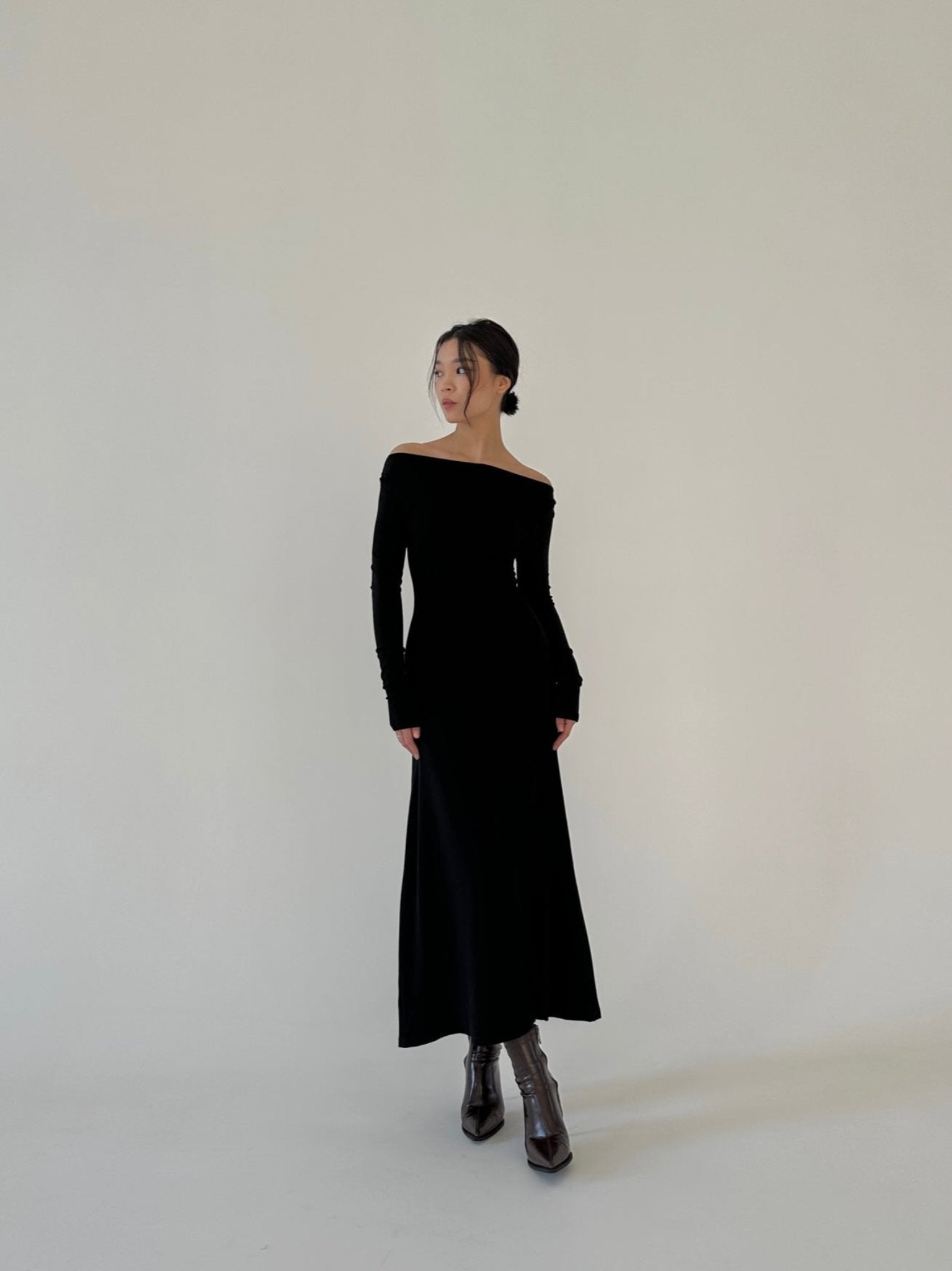 A woman stands in front of a plain white background, wearing the Sienna maxi dress, crafted from luxurious fabric. The long-sleeved black dress extends down to her ankles. Her hair is tied back and she pairs the ensemble with black pointed-toe boots. She gazes to her left with a relaxed posture.