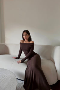 A woman with long dark hair is seated on a cream-colored sofa, wearing the Sienna maxi dress made of high-quality fabric. She looks directly at the camera as the versatile dress accentuates her figure against the minimal background of plain white walls.