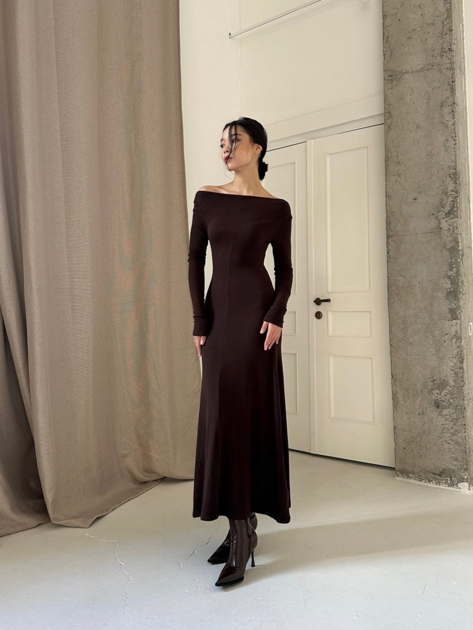 A woman stands in a minimalistic, modern room with a concrete pillar and beige curtains. She is wearing an elegant Sienna maxi dress and black pointed-toe boots. Her hair is styled up as she gazes to the side. The room has a white door and light-colored walls, enhancing the sophisticated ambiance.